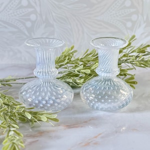 Op de afbeelding: Twee heldere glazen vaasjes met een gestructureerd, gestippeld patroon op de bolvormige basis en een gegroefde hals. De vazen hebben een delicaat ontwerp, ideaal voor kleine bloemen of groen. De achtergrond bevat groen.