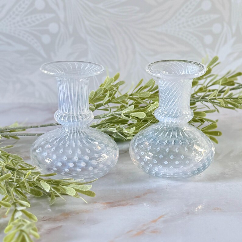 Op de afbeelding: Twee heldere glazen vaasjes met een gestructureerd, gestippeld patroon op de bolvormige basis en een gegroefde hals. De vazen hebben een delicaat ontwerp, ideaal voor kleine bloemen of groen. De achtergrond bevat groen.