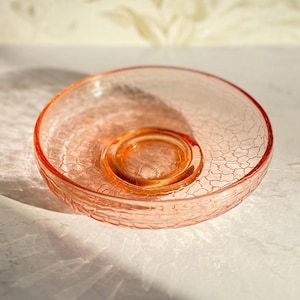 May include: A vintage pink glass bowl with a textured, crackled surface. The bowl has a circular shape with a raised center and a slightly flared rim. The glass is translucent, allowing light to pass through.