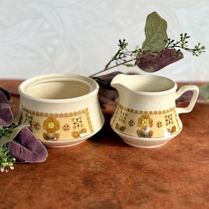 May include: A vintage cream-coloured sugar bowl and creamer set. Both pieces feature a decorative band with a repeating floral pattern in shades of yellow, brown, and green. The sugar bowl is open, and the creamer has a handle and spout.