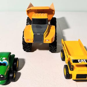 May include: Three toy construction vehicles: a green tractor, a large orange dump truck, and a smaller yellow dump truck with "CAT" printed on it. The tractor has a smiling face design. The dump trucks have black tires.