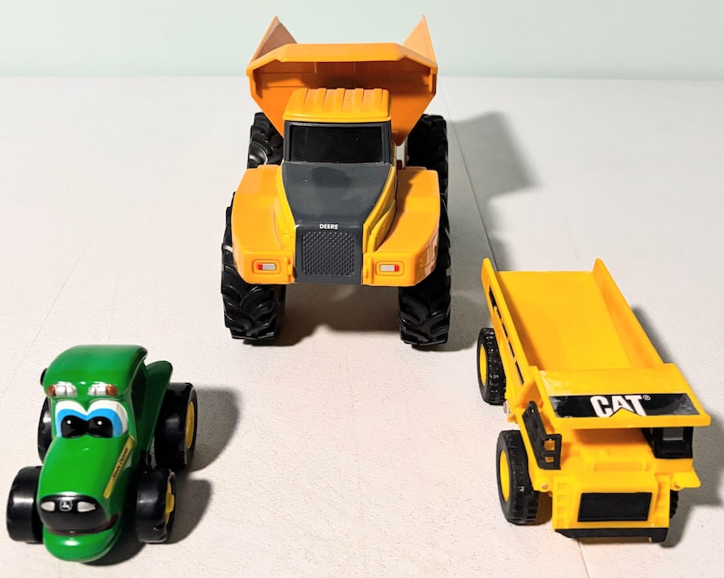May include: Three toy construction vehicles: a green tractor, a large orange dump truck, and a smaller yellow dump truck with "CAT" printed on it. The tractor has a smiling face design. The dump trucks have black tires.