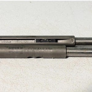 May include: A close-up of a metallic grey mechanical component. The component has a cylindrical shape with a rectangular section and a small brass fitting. The text "HEULE GOFA12 536" is visible on the side.