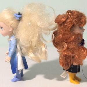 Op de afbeelding: Twee kleine poppen, een met blond haar en een blauwe jurk, de andere met rood haar en een donkere outfit. De poppen staan naast elkaar, naar links gericht. De blonde pop heeft een blauw haaraccessoire.