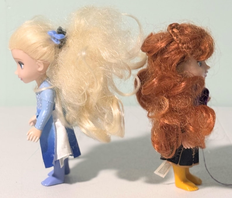 Op de afbeelding: Twee kleine poppen, een met blond haar en een blauwe jurk, de andere met rood haar en een donkere outfit. De poppen staan naast elkaar, naar links gericht. De blonde pop heeft een blauw haaraccessoire.