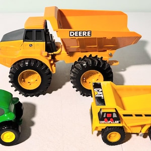 May include: Three toy construction vehicles: a large yellow dump truck with "DEERE" on the side, a small green tractor with "John Deere" on the side, and a yellow dump truck with "CAT" on the side.