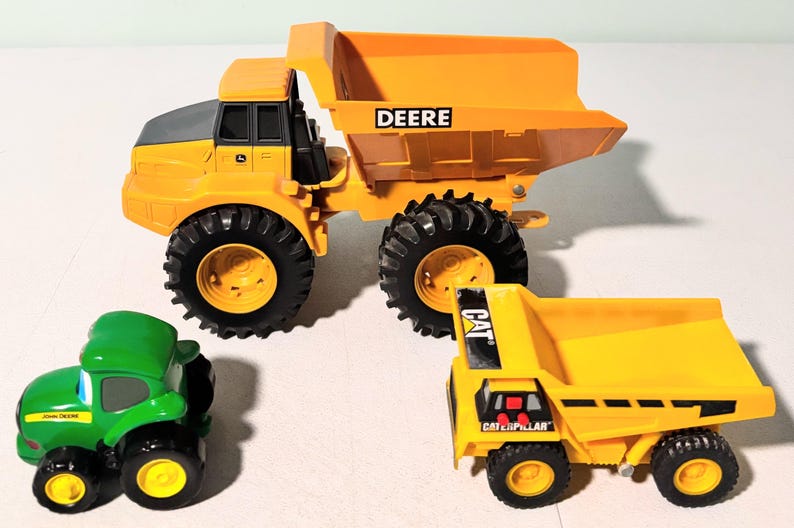 May include: Three toy construction vehicles: a large yellow dump truck with "DEERE" on the side, a small green tractor with "John Deere" on the side, and a yellow dump truck with "CAT" on the side.