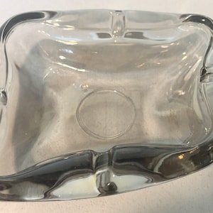 Puede incluir: Un cenicero de vidrio transparente con forma rectangular y esquinas redondeadas. El cenicero tiene cuatro apoyos para cigarrillos y una hendidura circular en el centro. El vidrio es transparente.