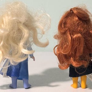 Op de afbeelding: Twee poppen van achteren gezien. De ene heeft lang, golvend, blond haar en een blauwe jurk. De andere heeft lang, krullend, rood haar, een zwarte rok en gele laarzen. Beide poppen hebben labels aan hun kleding.