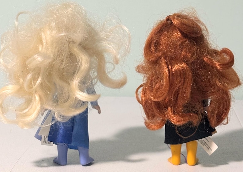 Op de afbeelding: Twee poppen van achteren gezien. De ene heeft lang, golvend, blond haar en een blauwe jurk. De andere heeft lang, krullend, rood haar, een zwarte rok en gele laarzen. Beide poppen hebben labels aan hun kleding.