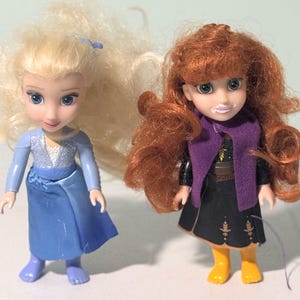 Op de afbeelding: Twee kleine poppen, een met blond haar in een blauwe jurk en de andere met rood haar in een zwarte jurk met een paarse sjaal. De poppen hebben blauwe ogen en staan rechtop. De pop met rood haar draagt gele laarzen.