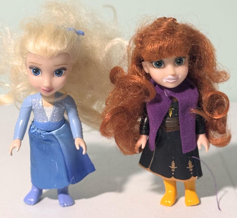 Op de afbeelding: Twee kleine poppen, een met blond haar in een blauwe jurk en de andere met rood haar in een zwarte jurk met een paarse sjaal. De poppen hebben blauwe ogen en staan rechtop. De pop met rood haar draagt gele laarzen.