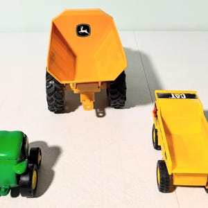May include: Three toy construction vehicles: a green tractor, a yellow dump truck with "CAT" text, and a large yellow dump trailer with a John Deere logo. The toys have black tires and are set against a white background.