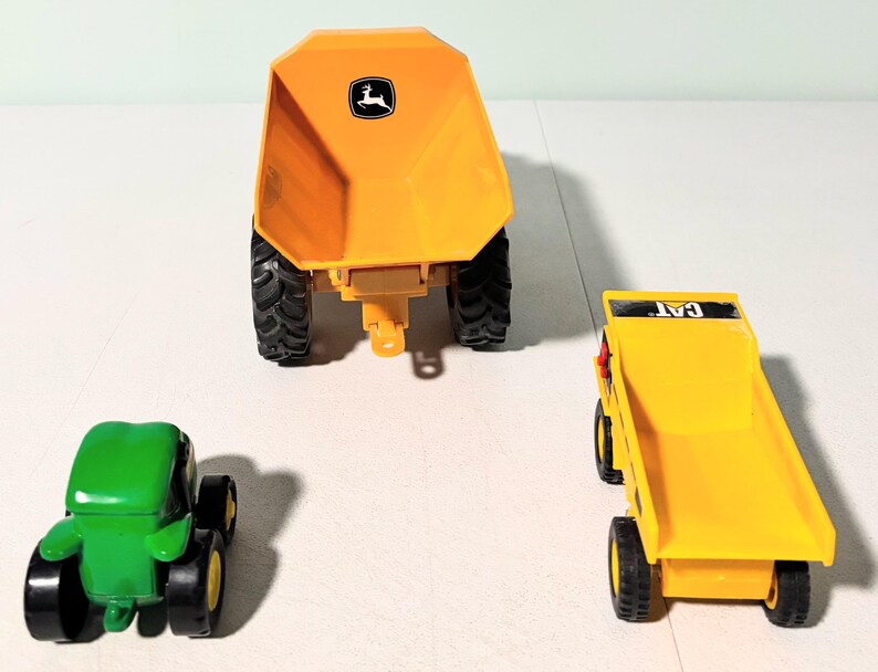 May include: Three toy construction vehicles: a green tractor, a yellow dump truck with "CAT" text, and a large yellow dump trailer with a John Deere logo. The toys have black tires and are set against a white background.