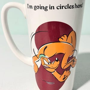 Peut inclure: Mug en céramique blanche avec une anse incurvée et un intérieur bordeaux. La tasse présente une image de dessin animé de Pluto, un chien orange, et le texte "I'm going in circles here!"