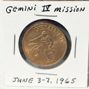 May include: A gold-colored coin commemorating the Gemini IV mission, featuring an astronaut and the mission name. The coin is encased in a white card with the text "Gemini IV mission" and "June 3-7, 1965" written in black ink.