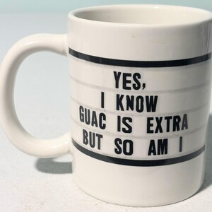 Puede incluir: Taza de cerámica blanca con asa curva. La taza presenta texto negro que dice "YES, I KNOW GUAC IS EXTRA BUT SO AM I" sobre un fondo a rayas. La taza tiene un diseño simple y limpio.