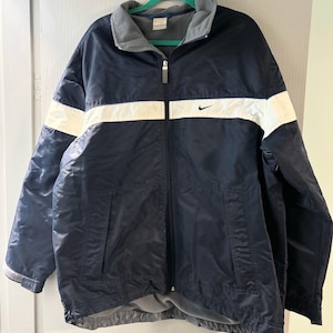 Może przedstawiać: Granatowa kurtka zapinana na zamek z białymi paskami na klatce piersiowej i rękawach. Posiada szary polarowy kołnierz i logo Nike na piersi. Swobodna, sportowa odzież wierzchnia.