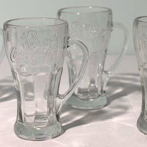 Könnte beinhalten: Vier transparente Glasbecher mit Henkeln. Die Becher haben ein klassisches Coca-Cola-Design, das in das Glas geätzt ist. Die Becher sind transparent und haben einen abgerundeten Boden und einen ausgestellten oberen Rand.
