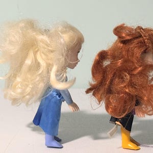 Op de afbeelding: Twee kleine poppen, een met blond haar en een blauwe jurk, de andere met rood haar en een zwarte outfit met gele laarzen. De poppen staan naast elkaar en tonen hun gedetailleerde kenmerken en levendige kleuren.