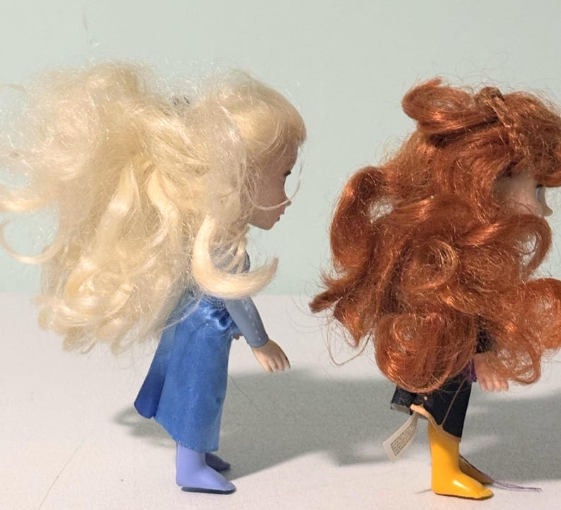 Op de afbeelding: Twee kleine poppen, een met blond haar en een blauwe jurk, de andere met rood haar en een zwarte outfit met gele laarzen. De poppen staan naast elkaar en tonen hun gedetailleerde kenmerken en levendige kleuren.