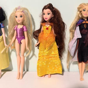 May include: Four dolls of animated characters. Snow White wears a blue top and yellow skirt. Rapunzel has long blonde hair and a pink outfit. Belle wears a yellow gown. Anna wears a black dress with a purple cape.