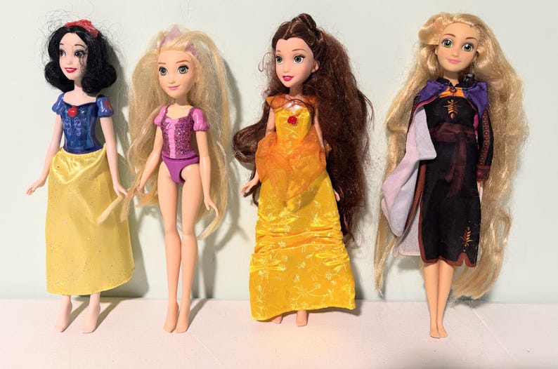 May include: Four dolls of animated characters. Snow White wears a blue top and yellow skirt. Rapunzel has long blonde hair and a pink outfit. Belle wears a yellow gown. Anna wears a black dress with a purple cape.