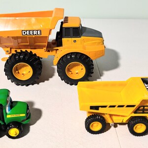 May include: Three toy construction vehicles: a large yellow dump truck with "DEERE" on the side, a smaller yellow dump truck with "CAT" on the side, and a green tractor with "JOHN DEERE" on the side. The vehicles have black tires.