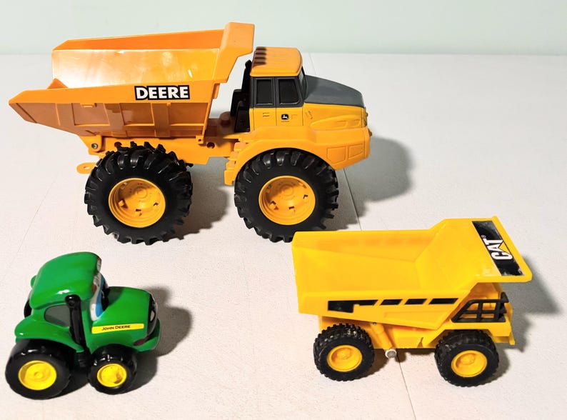 May include: Three toy construction vehicles: a large yellow dump truck with "DEERE" on the side, a smaller yellow dump truck with "CAT" on the side, and a green tractor with "JOHN DEERE" on the side. The vehicles have black tires.