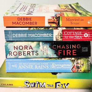 Op de afbeelding: Een stapel van vijf boeken met kleurrijke covers. Titels zijn onder meer "Married in Texas", "Cottage by the Sea" en "Chasing Fire". Auteurs zijn Debbie Macomber, Nora Roberts en Annie Rains. De boeken zijn op elkaar gestapeld.