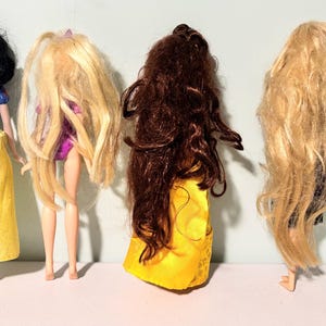 May include: Four dolls are displayed, each with distinct hairstyles and outfits. One doll has black hair and a yellow skirt, while the others have long blonde and brown hair. The dolls are standing in a row, showcasing their backs.