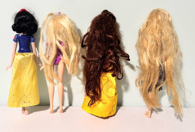 May include: Four dolls are displayed, each with distinct hairstyles and outfits. One doll has black hair and a yellow skirt, while the others have long blonde and brown hair. The dolls are standing in a row, showcasing their backs.