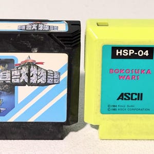 May include: Two vintage video game cartridges. One is black with a colorful label featuring a cartoon warrior. The other is yellow with a blue label and cartoon characters. Both are retro gaming collectibles.