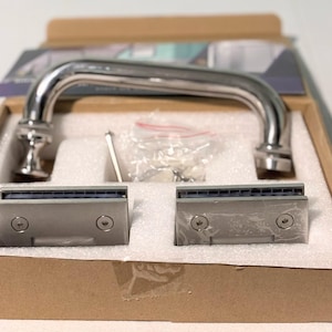 May include: A stainless steel shower door handle and two matching hinges are displayed in a cardboard box. The handle is curved and has decorative ends. The hinges are rectangular with a brushed metal finish. Hardware is included.