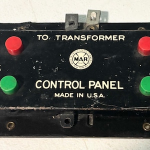 Può includere: Un pannello di controllo nero vintage con pulsanti rossi e verdi, etichettato "TO TRANSFORMER" e "CONTROL PANEL MADE IN U.S.A." Il pannello ha connettori metallici ed è probabilmente per un trenino o un dispositivo elettrico simile.