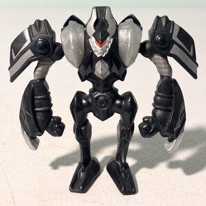 Peut inclure: Figurine de robot noir et argenté avec des bras et des jambes articulés. Le jouet présente un design détaillé avec des accents argentés sur les épaules, les bras et la tête. Le visage du robot comporte un détail rouge.