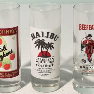 以下が含まれることがあります： ストリ・ラズベリーウォッカ、マリブカリビアンホワイトラムココナッツ、ビーフィーターロンドンドライジンのラベルが付いた3つの透明なショットグラス。ストリのグラスにはラズベリーが描かれています。マリブのグラスにはヤシの木があります。ビーフィーターのグラスには赤い服を着た人物がいます。