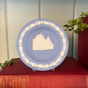 May include: A light blue decorative plate with a white relief of a building and the word "Harrods". The plate has a white floral border. It is displayed on a clear stand, resting on two red books.