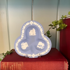 May include: A light blue ceramic ashtray with white relief designs and a floral border. The ashtray is clover-shaped and rests on a clear stand, placed on top of two red books with gold lettering.
