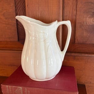 May include: A cream-colored ceramic pitcher featuring a fluted, vertical ribbed body, a curved handle, and a scalloped rim. The pitcher has a glossy finish and a molded decorative pattern near the top, showcasing a classic, elegant design.