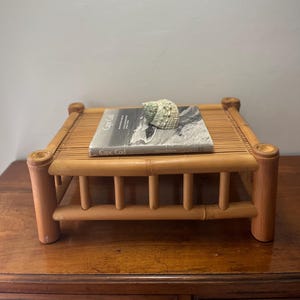 May include: A small, square bamboo table with rounded corner posts. A book titled "Cape Cod" and a light green seashell rest on the table's slatted top. The table is on a wooden surface, with a neutral wall in the background.