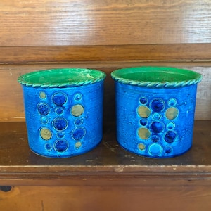 May include: Two cylindrical ceramic planters featuring a vibrant blue glaze with a textured surface. Each planter is decorated with a pattern of raised circular motifs in shades of dark blue and gold, finished with a green rope-style rim.