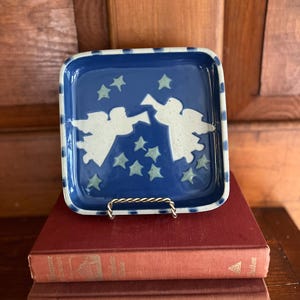 May include: A square, ceramic dish with a blue background and white angel and star designs. The dish has a blue and white dotted border. It is displayed on a small metal stand, resting on top of two red books.