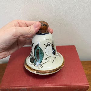 May include: A ceramic bell with a bird and floral design. The bell is cream-colored with brown, teal, and black accents. The bell is held by a hand and sits on top of two red books.