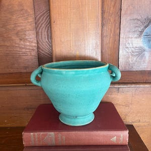 May include: A turquoise ceramic vase with two handles, sitting atop two antique books with red covers. The vase has a wide opening and a pedestal base. The background is a wooden surface.