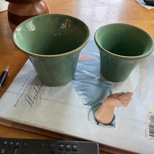 May include: Two sage green ceramic pots with flared rims, one larger than the other, resting on a book titled The Best of Martha Stewart Living Weddings. A black pen and a remote control are visible nearby on the wooden surface.