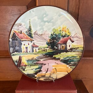May include: A decorative ceramic plate featuring a hand-painted landscape scene with houses, trees, and a stream. The plate has a brown rim and is displayed on a metal stand, resting on a stack of red books. The artwork includes shades of green, yellow, and brown.