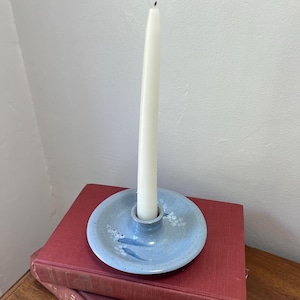 May include: A light blue ceramic candle holder with white floral accents and a white taper candle. The candle holder is sitting on top of two red books. The candle is approximately 10 inches tall.