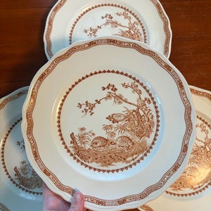 May include: A set of four white ceramic dinner plates with scalloped edges and a brown transferware pattern. The central design features two birds resting in a landscape with trees and foliage, surrounded by a decorative geometric border.