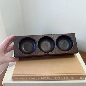 May include: A rectangular dark brown wooden block featuring three circular carved indentations arranged in a horizontal row. The block is positioned atop a tan book titled Martha Stewart's New Old House, which rests on a white book.